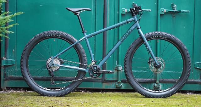 A rigid mountain bike with fat tyres leaning against a turquoise lock-up, The bike is blue and has the Genesis logo on it