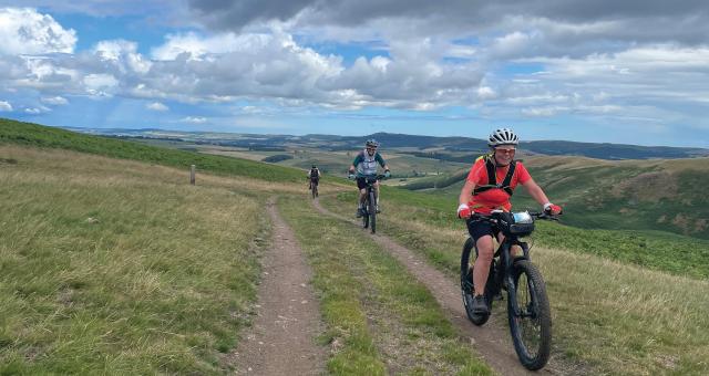 Three women are cycling on mountain bikes along an off-road track through hills in Northumberland. They are wearing MTB clothing, cycling sunglasses and helmets.