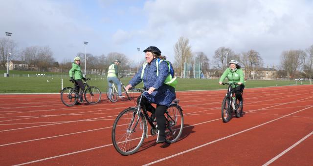 One white male and three white females are cycling along an athletics track. They have high-vis jackets on, and are wearing helmets and beaming smiles as they ride past the camera