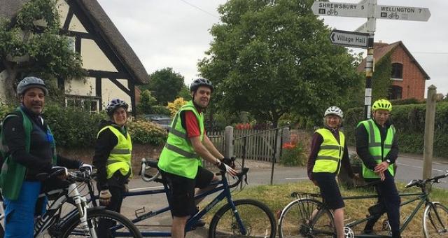 Jean Breakell is sat in the stoker position (at the back) on the tandem under a street signpost in a village. There are other cyclists around her, all wearing high vis vests