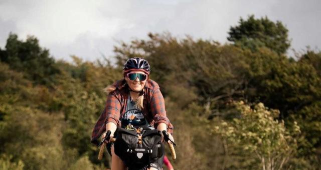 Jo Lankester is cycling along a wide flat grassy track wearing cycling glasses and a red checked shirt. There is a woodland behind her