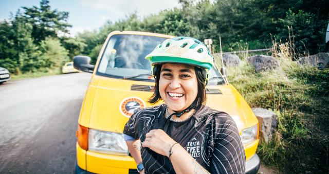 Jo Shwe is smiling wearing a green cycle helmet and standing in front of a yellow van which is parked on the side of a tree lined road
