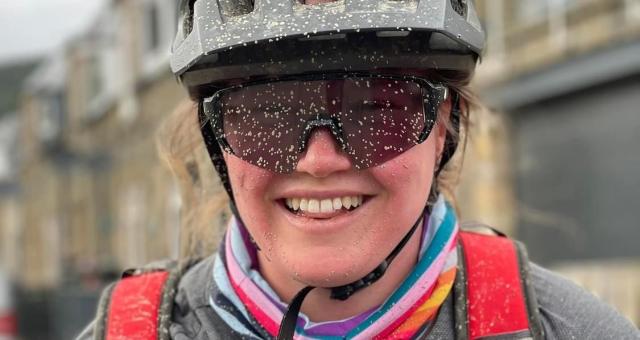 Jodie Brumhead is pictured wearing a black helmet and sunglasses. She is smiling and her cheeks are flushed and there are small flecks of mud covering her face and top