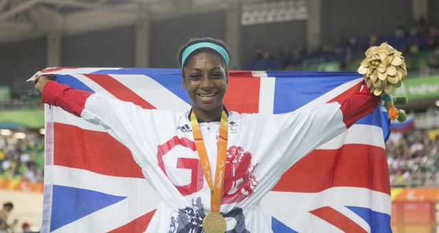 Kadeena Cox is holding the Union Jack flag behind her and is wearing a gold medal that has the words Rio 2016 along the ribbon. She is also holding gold flowers and is pictured inside a stadium