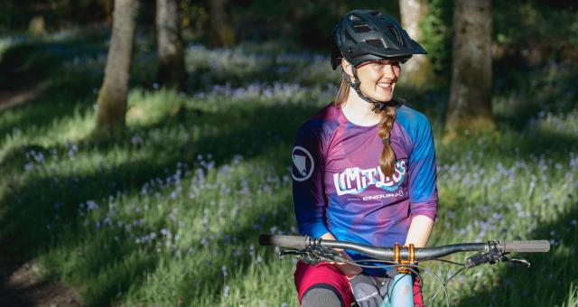 Katie May is sitting astride a mountain bike in a woodland. She is wearing a helmet and a Limitlass cycling jersey. Her blonde hair is in a plait. She is smiling.