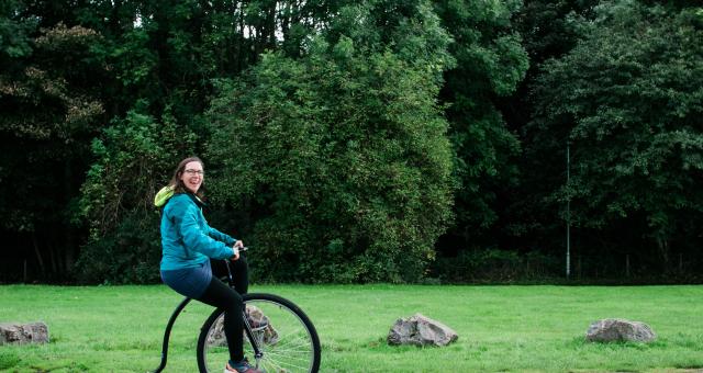 Katie Noble is smiling as she rides a penny farthing along a path. Behind her there is a grass lawn lined with trees