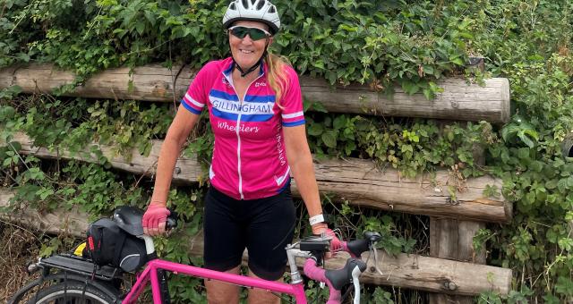 Mary Emerson-Reed is standing with a bright pink touring bike. She is wearing black shorts and a matching bright pink Gillingham Wheelers short-sleeved jersey. She has a helmet and cycling sunglasses. She is smiling at the camera.