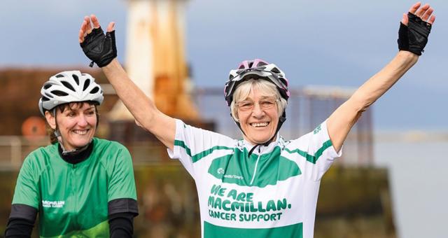 Mavis Paterson pictured at the finish line after completing her record-breaking LEJOG ride. She is smiling broadly and has both arms raised in the air. She is wearing a green t-shirt that reads ‘we are Macmillan Cancer Support’
