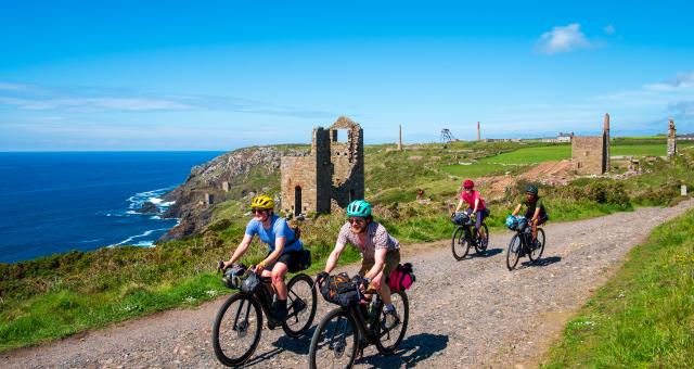 Cyclists riding along a coastal gravel trail.  There are ruins and the sea in the background.