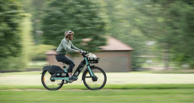 A woman is riding an e-bike in a park. It's green and black and has a Making cycling e-asier logo on it. There's a basket on the front with some shopping in it. The rider is wearing normal clothes and a cycle helmet. She is smiling