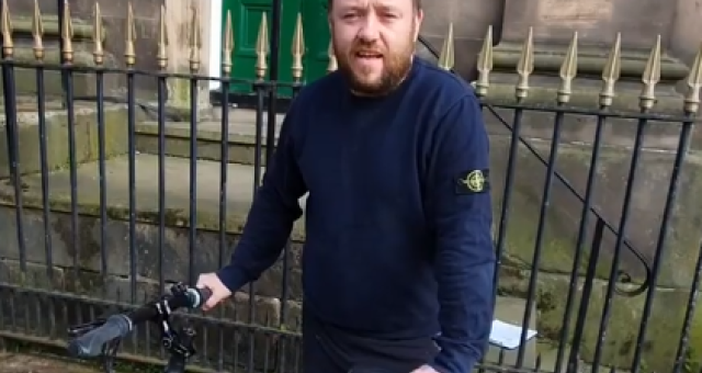 Freezeframe from a video of a man in a navy pullover standing behind a bicycle