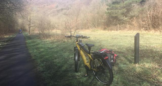 A bright yellow e-bike stands on the grass next to an off-road cycle route