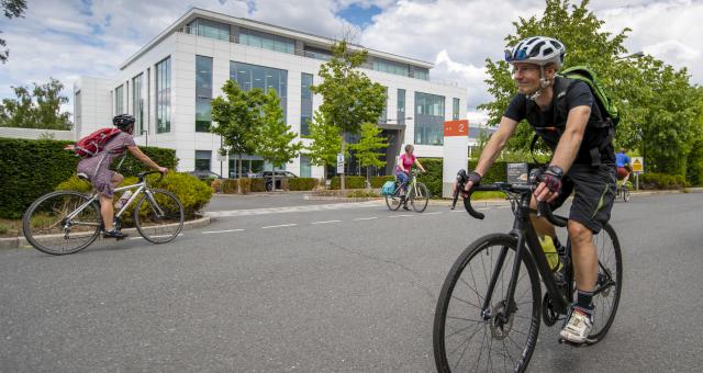 Man wearing helmet cycles towards camera while people cycle in opposite direction on other side of the road. Large white building, cloudy blue sky and trees in background.