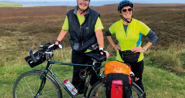 Two women stand behind a Dawes Galaxy touring bike that's loaded with panniers. They are both wearing helmets and high vis tops and smiling broadly at the camera. In the background is a coastline covered in heather.
