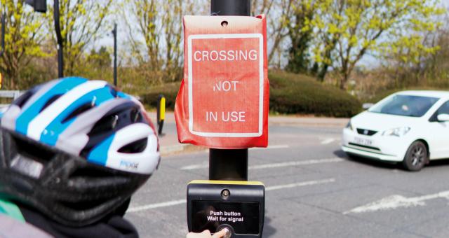 A woman goes to press the button for a pedestrian crossing but the lights have a cover over them which reads 'Crossing not in use'. A car approaches in the background