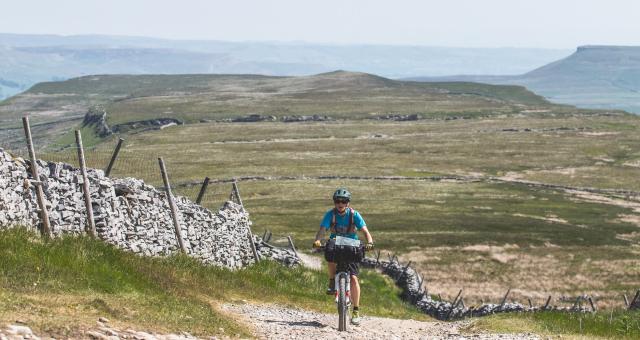 Woman cycles up a rough gravel track towards the camera, with an expansive moorland view behind her