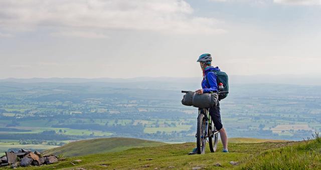 A cyclist standing over their bike, looking back over distant hills.