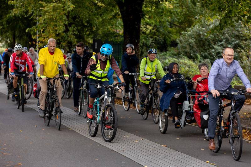 A large group of people are cycling on a path together. They are on a range of bikes, including adaptive, and wearing normal clothes. Some have helmets, some don't.