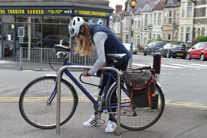 A woman is locking her blue Raleigh hybrid bike to a cycle hoop in an urban street. She is wearing trainers, blue jeans, blue body warmer and blue and white stripped top and a white cycling helmet.