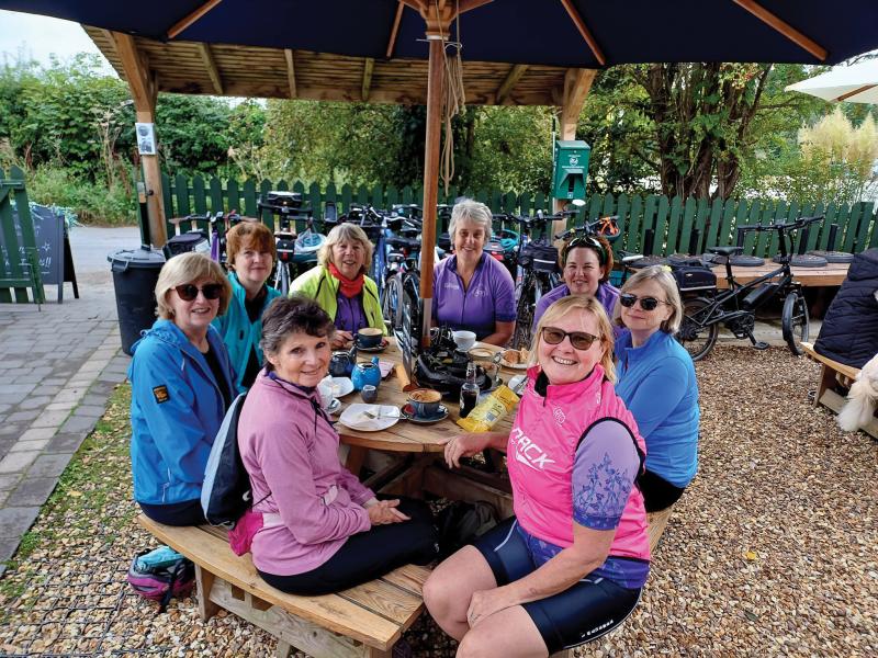 A group of women are sitting around a picnic table at a cafe. There's a selection of drinks and snacks in front of them. They're wearing cycling kit and their bikes can be seen leaning against a fence in the background. They are smiling at the camera.