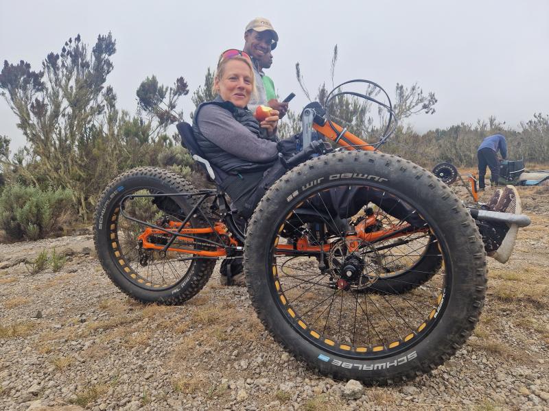 A woman is on a fat bike mountain trike. She's eating an apple and smiling at the camera. Two men are behind her.