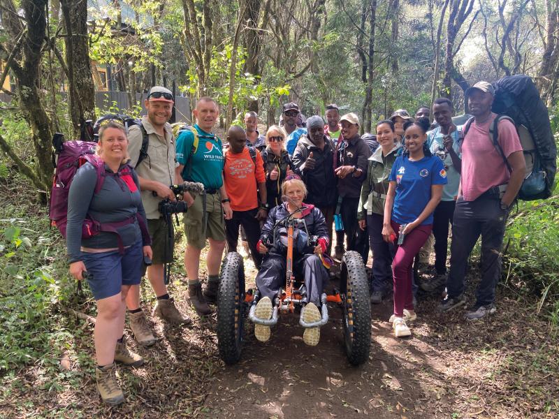 A mixed group of people is standing on a forest track. They have rucksacks and are wearing hiking kit. In the middle of the group is a woman on a mountain trike