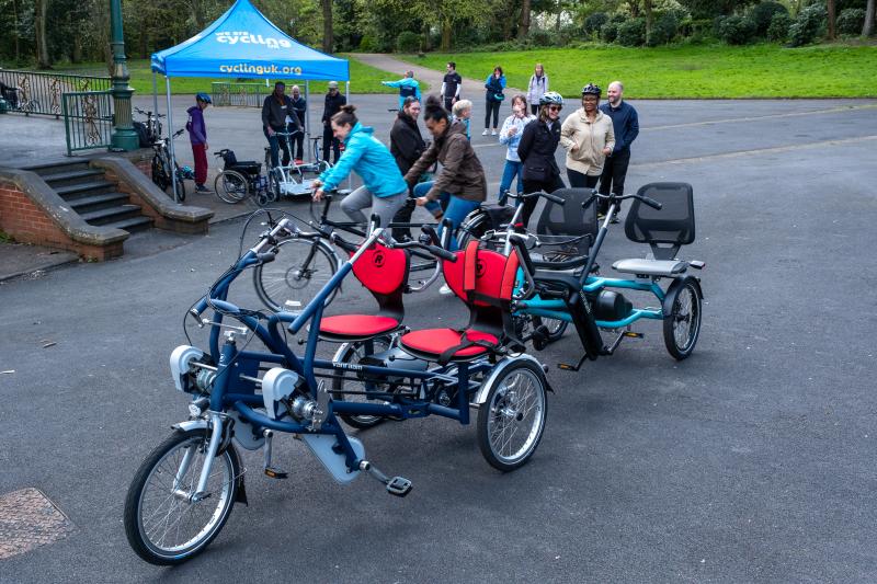 Two access trikes sitting in the foreground in a park in Manchester with Cycling UK staff and members of the public standing behind them talking ©2024 iD8 Photography
