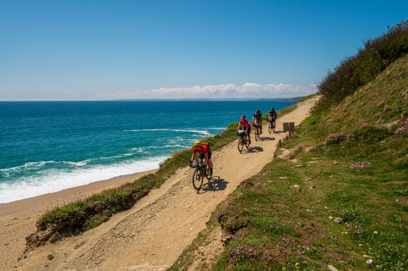 Four people cycling downhill on an off road route with hills to their left and the sea to their right on a sunny day