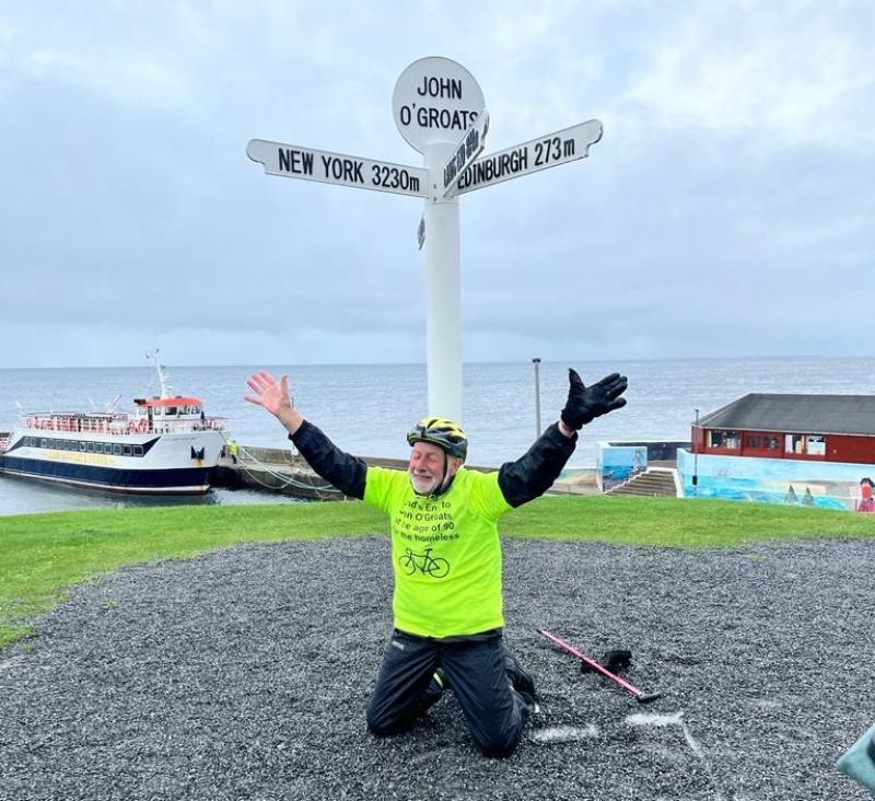 A man kneeling on the ground under the signpost at John o’ Groats