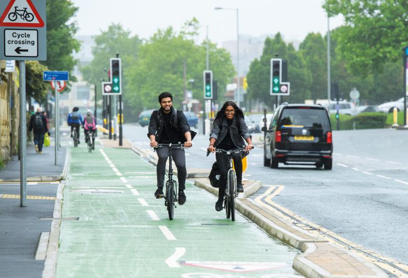 Two people are cycling along a green segregated cycle path with a pavement to one side and a road to the other. One is on a folding bike, the other a hybrid. They are wearing normal clothes. It's raining, but they're smiling