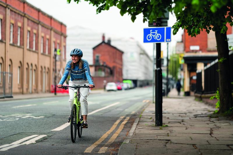 A young woman cycles along a quiet urban street, she is wearing casual clothes and a helmet and is smiling away from the camera