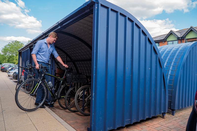 A man is wheeling his bike into a large blue bike shed with lots of other bikes already in there