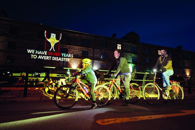 Two men and a young girl cycle on bikes in front of a lit up building