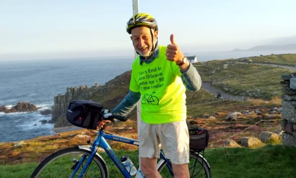 A man stands with a bike under the Land’s End signpost. He is wearing a bright yellow T-shirt and giving a thumbs up to the camera