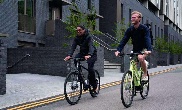 Two men are riding e-cycles side by side along an urban street. They are wearing normal clothes with no cycling helmet.