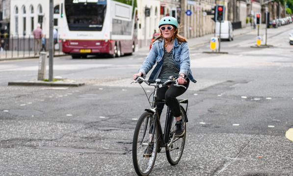 A woman is riding a black hybrid bike with a rear rack on it on an urban street. She is wearing black trousers, a stripy top and blue denim jacket. She has long red hair, a turquoise cycling helmet and sunglasses.
