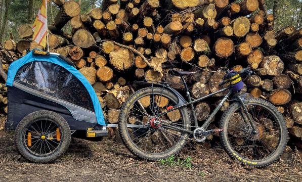 A black Surly mountain bike with a blue and black child trailer attached is leaning against a big log pile in a forest