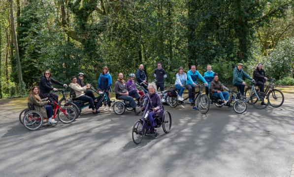 A group of adults in a park on a variety of non-standard cycles, tandems, e-assist cycles, handcycles and tricycles