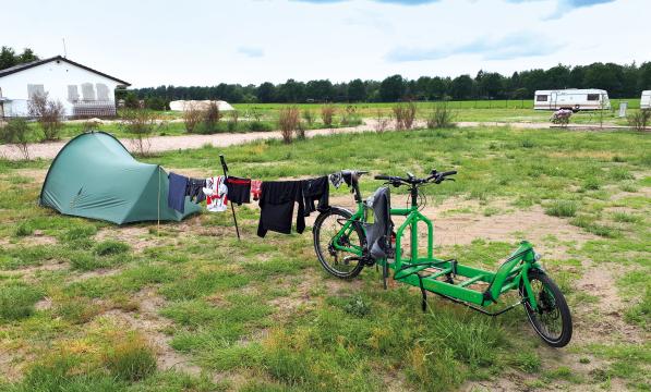 A tent sits on a grassy campsite, with a washing line of cycling clothes hung between it and a bright green cargo bike