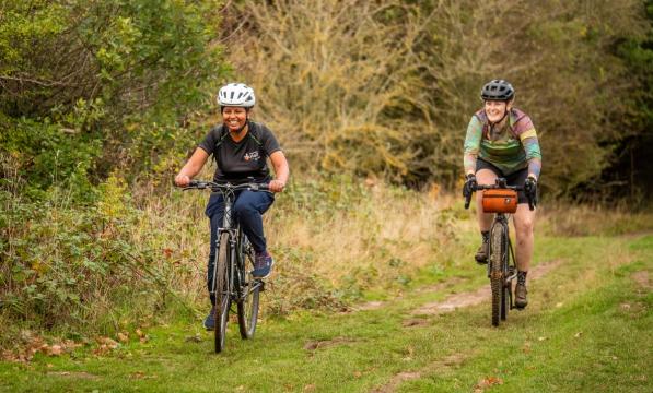Two women are riding along a grassy path in the countryside. One is riding a hybrid bike, the other a gravel bike. They are quite muddy and are laughing
