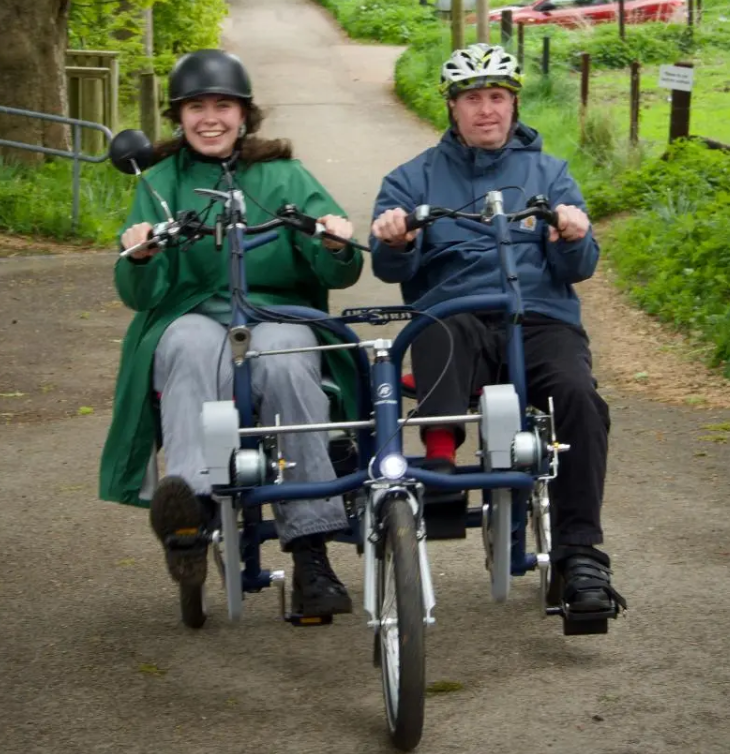 Two people ride side by side on a blue tandem tricycle along a country lane, both wearing helmets and smiling