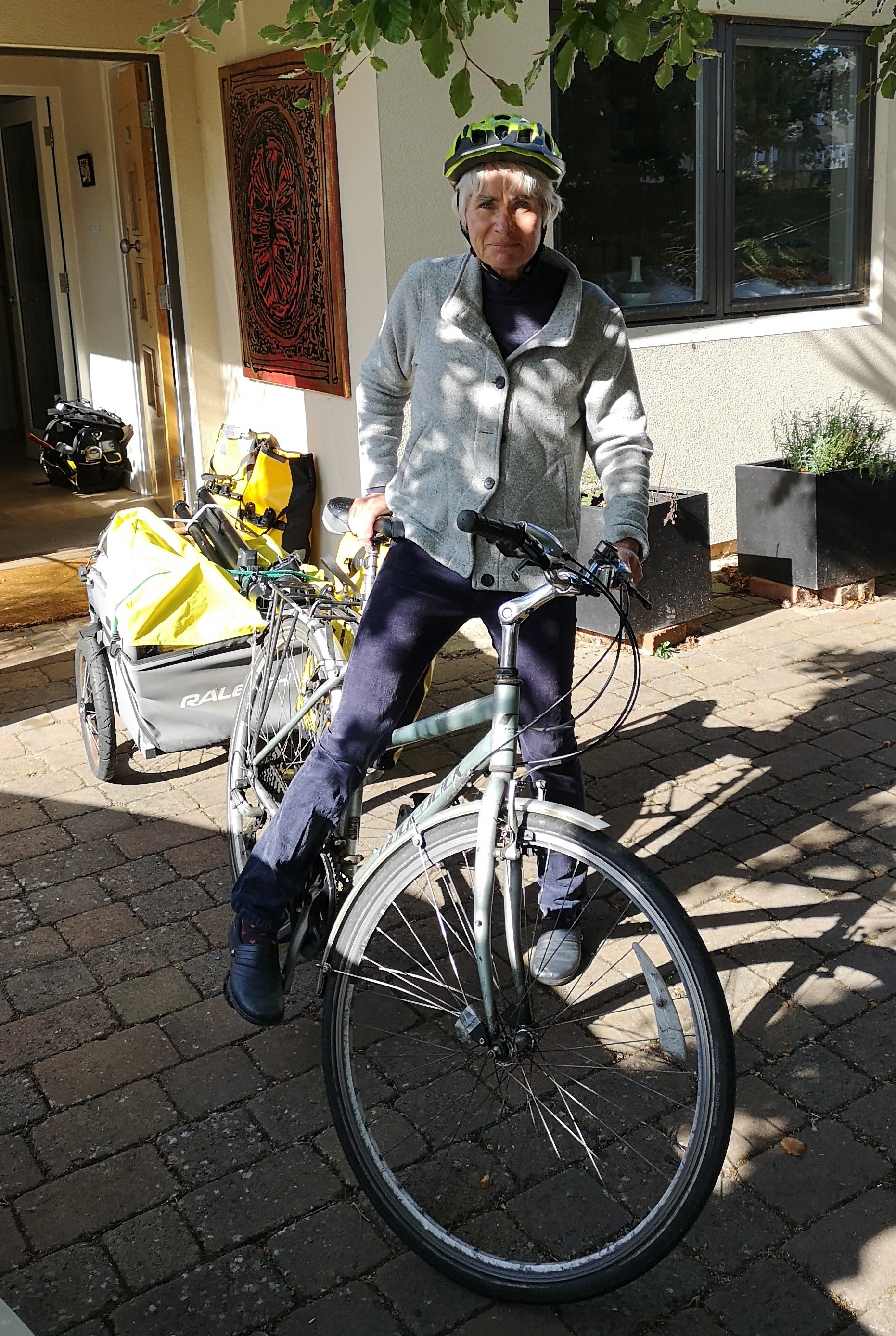 A woman is standing astride a grey hybrid bike with a packed trailer attached to it. She is wearing normal clothes: blue trousers and shoes and a grey fleece, as well as a cycle helmet.