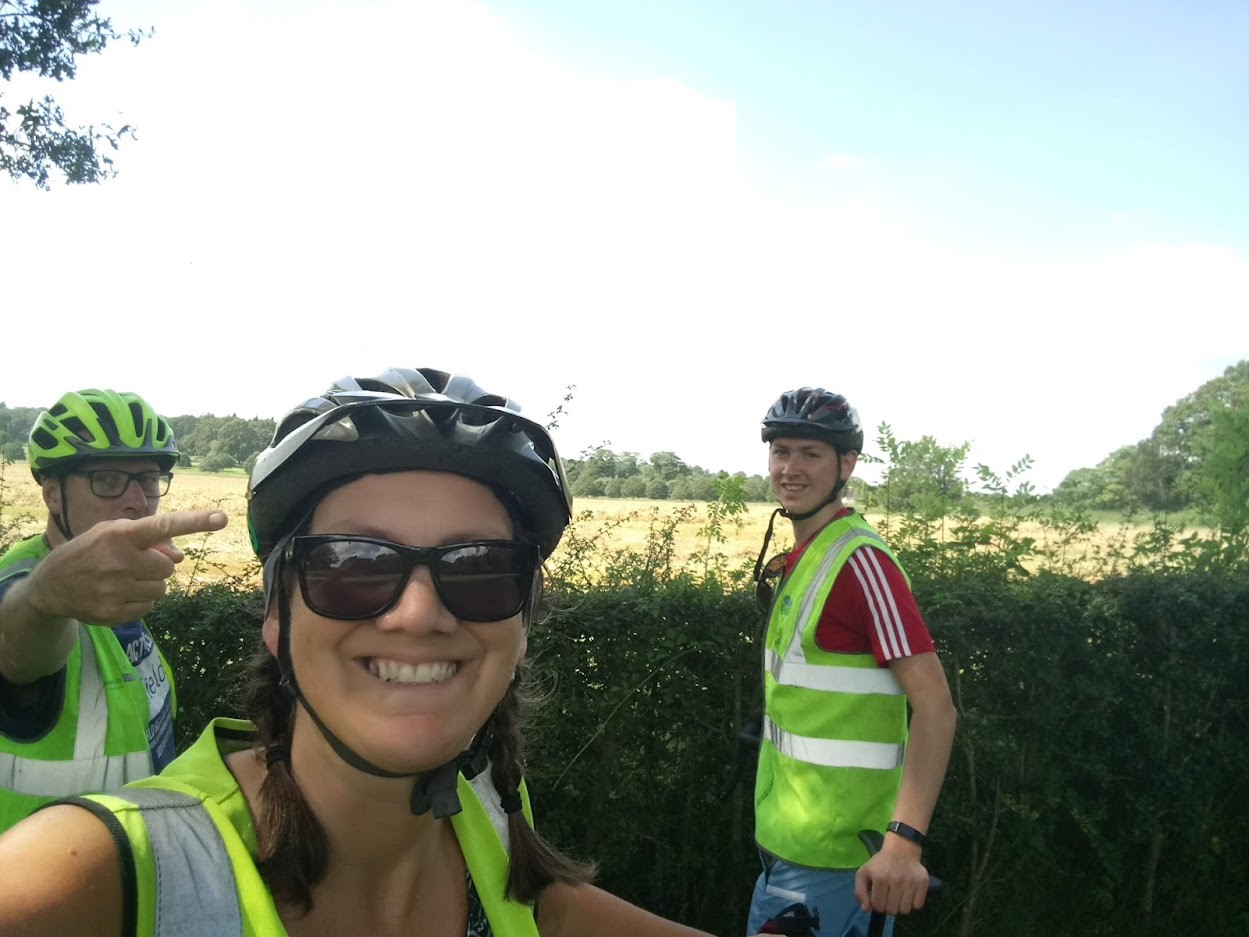 A selfie of three cyclists next to a hedge. The photo has been taken by a woman and behind her are two men, one to the left and one to the right. They are all wearing hi-vis jackets and cycling helmets