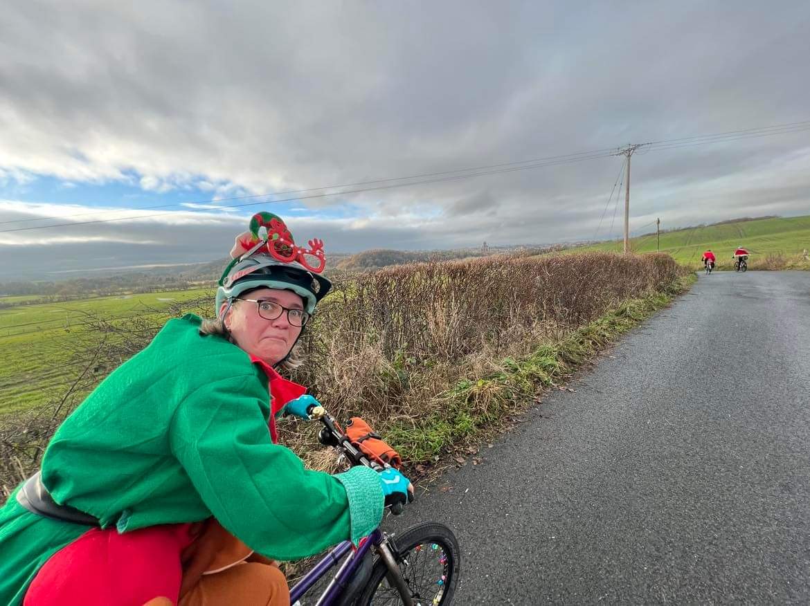 A woman is riding a bike while wearing a Christmas elf outfit. She is looking back at the camera and pulling a funny face