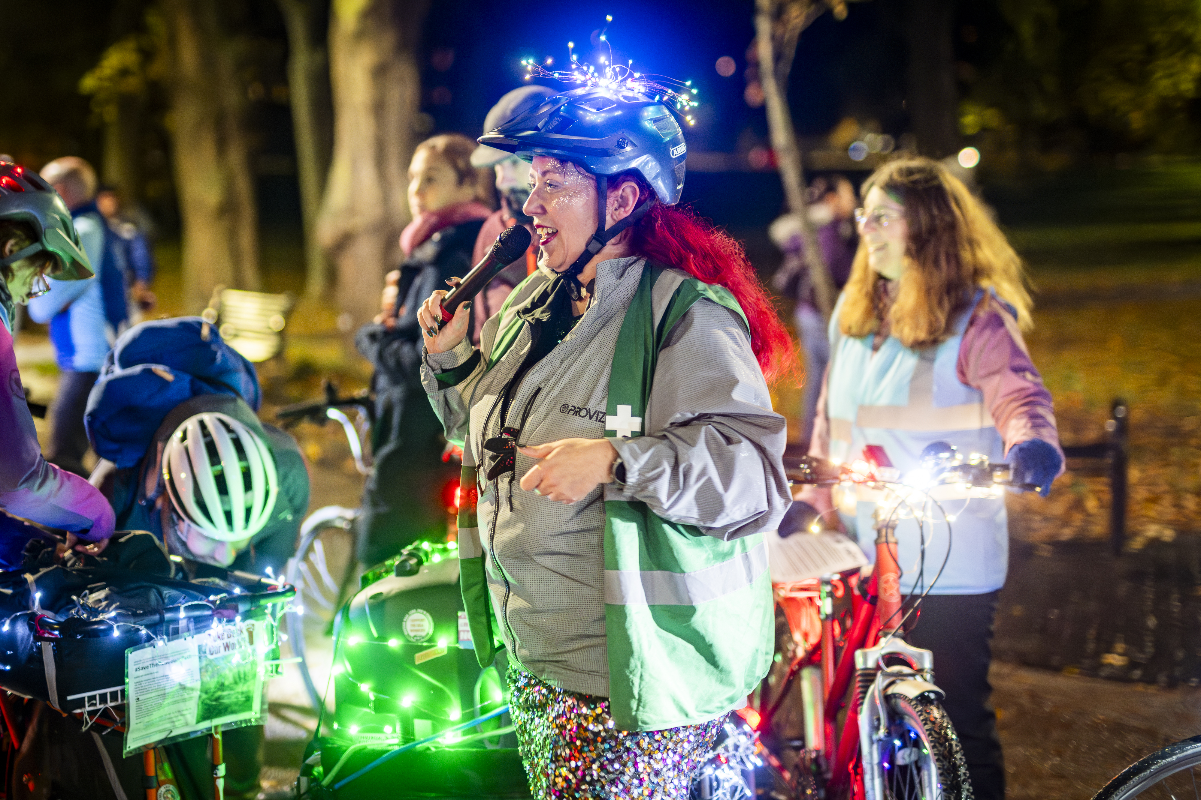 A woman speaks into a microphone at a mass women's evening ride. Her bike and helmet are decorated with fairy lights and she is wearing a sparkly skirt.