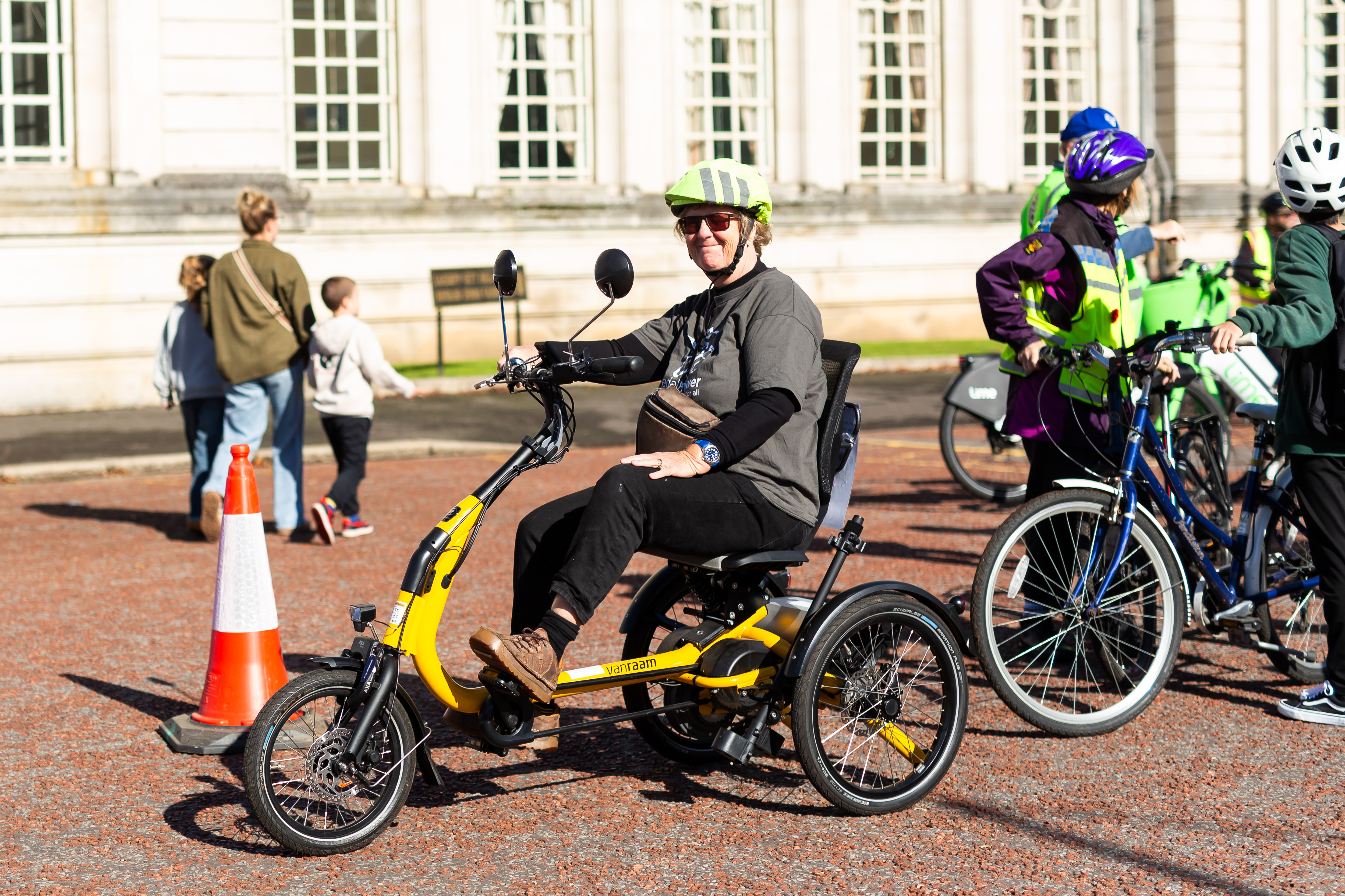 A woman is sitting on a bright yellow trike and smiling at the camera. There are more people on bikes and some people walking in the background