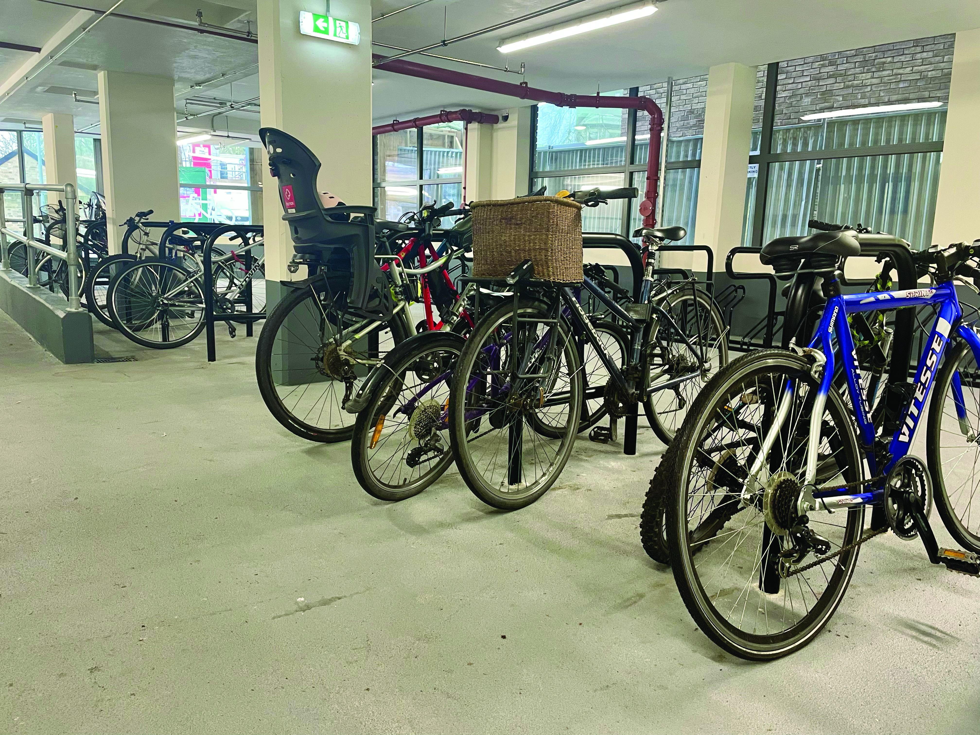 A variety of different bikes, including road bikes, hybrids, bikes with baskets, bikes with child seats, are all locked up in an undercover cycle park