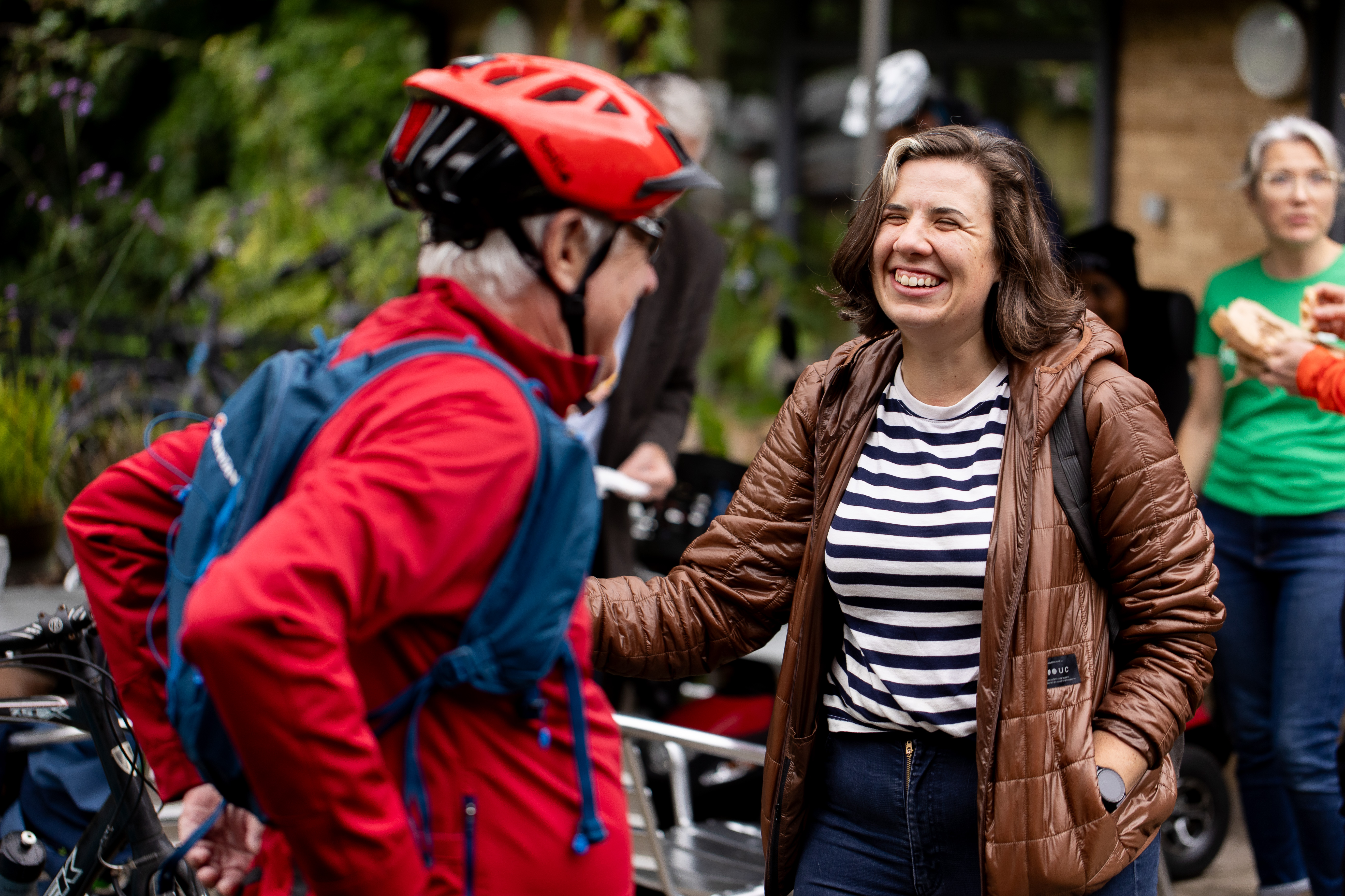 Two people are having a conversation. One is wearing a cycle helmet and jacket, the other is wearing normal clothes. They are laughing