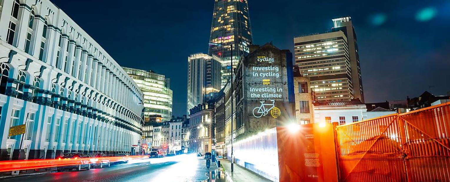 Night time photograph: the Cycling UK logo and a slogan saying "This machine fights climate change" are projected on the side of a building in London. The Shard skyscraper is in the background.