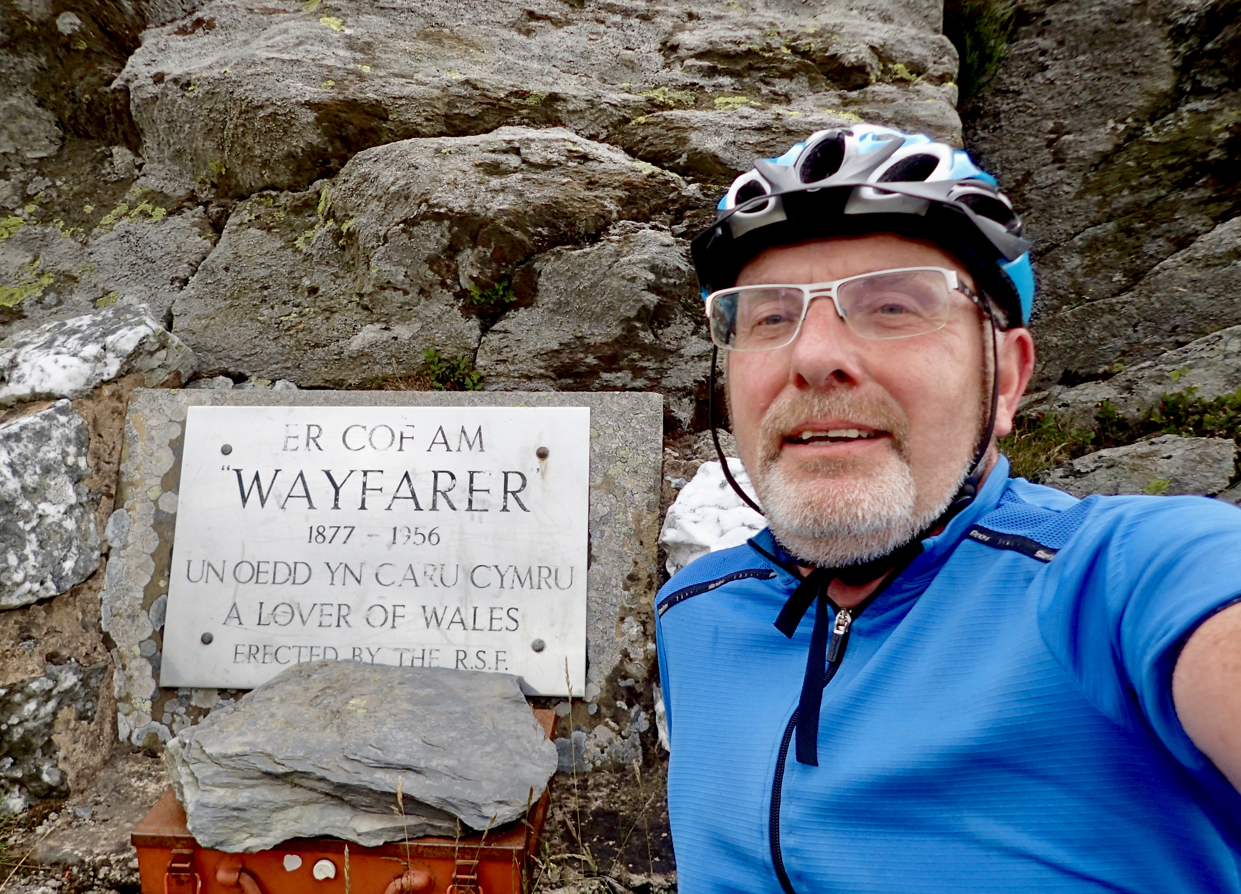 A man wearing a helmet clear glasses, and blue jacket poses in front of a silver plaque set into stone. The plaque reads: ER COF AM "Wayfarer" 1877-1956 UN OEDD YN CARU CYMRU A LOVER OF WALES Erected by the R.S.F